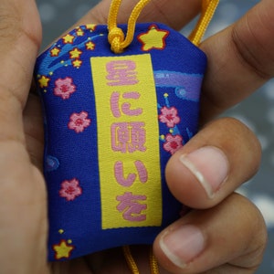 Könnte beinhalten: Ein blauer Stoffanhänger mit einem gelben Rechteck, das den japanischen Text "星に願いを" (bedeutet "Wünsche dir etwas vom Stern") enthält. Der Anhänger ist mit rosa Kirschblüten und gelben Sternen verziert.