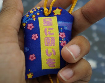 Puede incluir: Un amuleto de tela azul con un rectángulo amarillo que contiene el texto japonés "星に願いを" (que significa "Desear una estrella"). El amuleto está decorado con flores de cerezo rosas y estrellas amarillas.