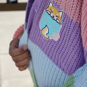 May include: A multi-colored knitted cardigan with a light pink, purple, light blue, and green color scheme. The cardigan has a cute cartoon fox patch on the front.