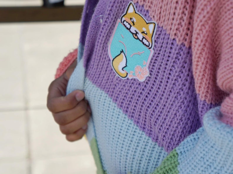 May include: A multi-colored knitted cardigan with a light pink, purple, light blue, and green color scheme. The cardigan has a cute cartoon fox patch on the front.