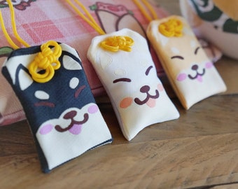 May include: Three small fabric pouches with embroidered dog faces. Each pouch has a yellow string tied around it with a knot at the top. The pouches are on a pink and white checkered background.