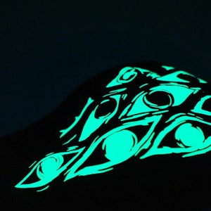 May include: Black garment featuring a glowing green eye design. The design consists of multiple stylized eyes, creating a striking visual effect against the dark background. The artwork has a hand-drawn aesthetic.