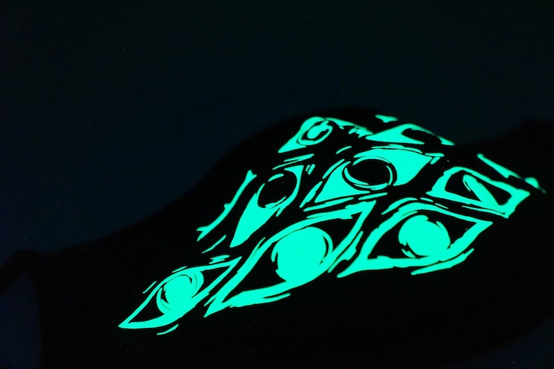 May include: Black garment featuring a glowing green eye design. The design consists of multiple stylized eyes, creating a striking visual effect against the dark background. The artwork has a hand-drawn aesthetic.