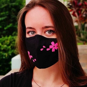 Könnte beinhalten: Eine Frau trägt eine schwarze Gesichtsmaske mit rosafarbenen Kirschblütenmustern. Die Maske bedeckt ihre Nase und ihren Mund.