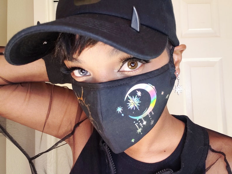 May include: A person wearing a black face mask with a crescent moon and stars design. The mask has a rainbow iridescent finish. The person is also wearing a black baseball cap and a black shirt.