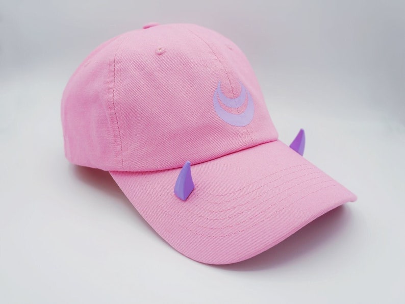 May include: A pink baseball cap with a purple crescent moon design and purple horns on the sides.