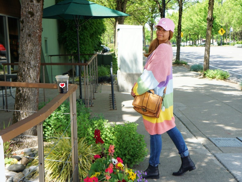 May include: A woman wearing a rainbow striped cardigan, a pink baseball cap, blue jeans, and black ankle boots. She is carrying a brown leather satchel bag.