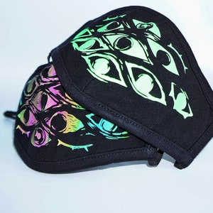 May include: Two black face masks with a repeating eye design in iridescent colors. The masks are made of a dark fabric and have a curved shape, with a black trim around the edges. The eye design has a neon green, pink, and blue color scheme.
