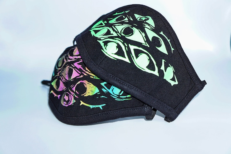 May include: Two black face masks with a repeating eye design in iridescent colors. The masks are made of a dark fabric and have a curved shape, with a black trim around the edges. The eye design has a neon green, pink, and blue color scheme.