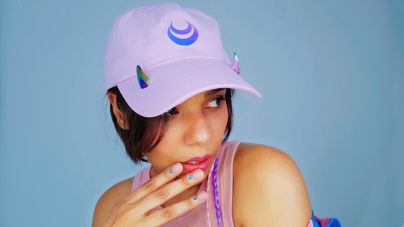 May include: A pink baseball cap with a blue and white crescent moon design. The cap has iridescent triangle shaped embellishments on the sides.