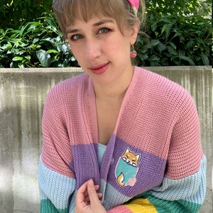 May include: A multi-colored striped cardigan with a light pink, purple, light blue, green, and yellow color scheme. The cardigan features a small embroidered patch of a Shiba Inu dog on the left side of the chest.