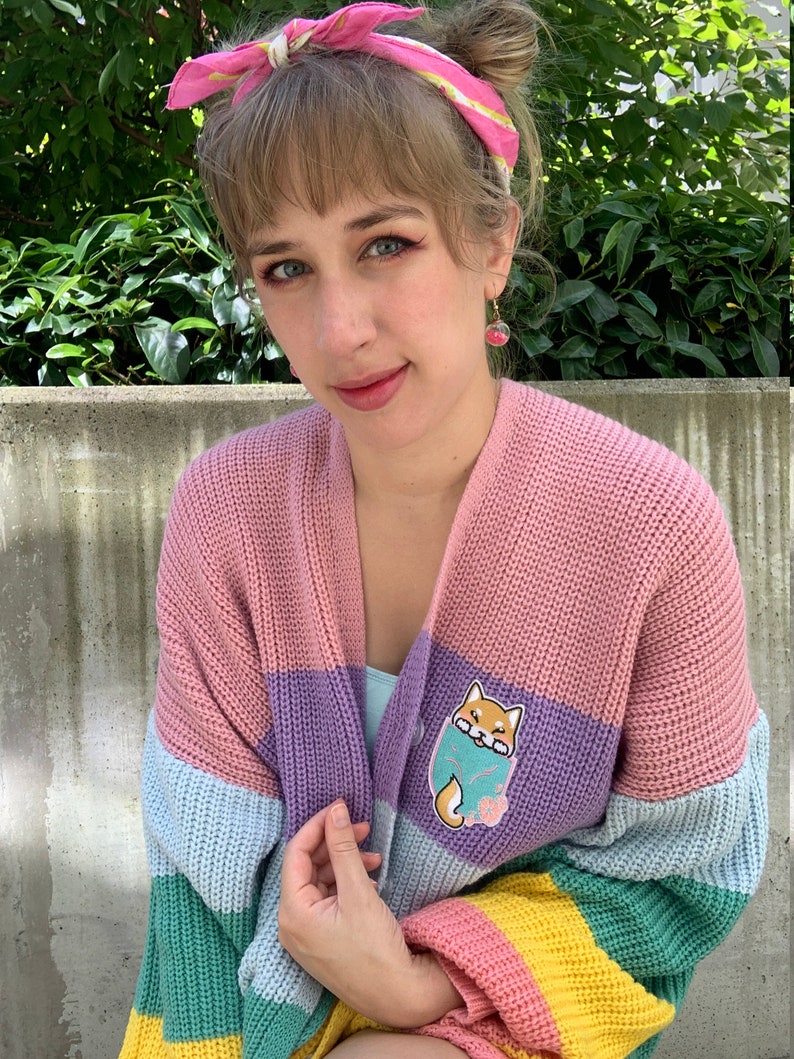 May include: A multi-colored striped cardigan with a light pink, purple, light blue, green, and yellow color scheme. The cardigan features a small embroidered patch of a Shiba Inu dog on the left side of the chest.