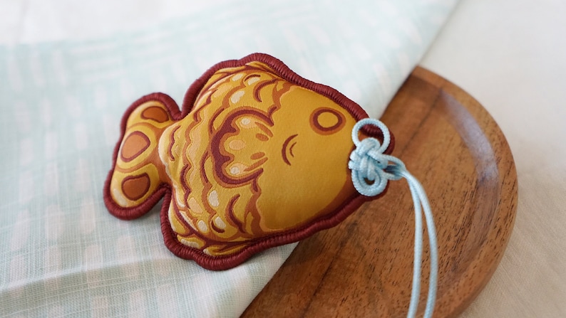 There is an Omamori protection charm that is shaped like a Taiyaki fish treat on a table cloth and a wooden plate. Blue strings are coming out of the Taiyaki's mouth in a Chinese fortune knot.