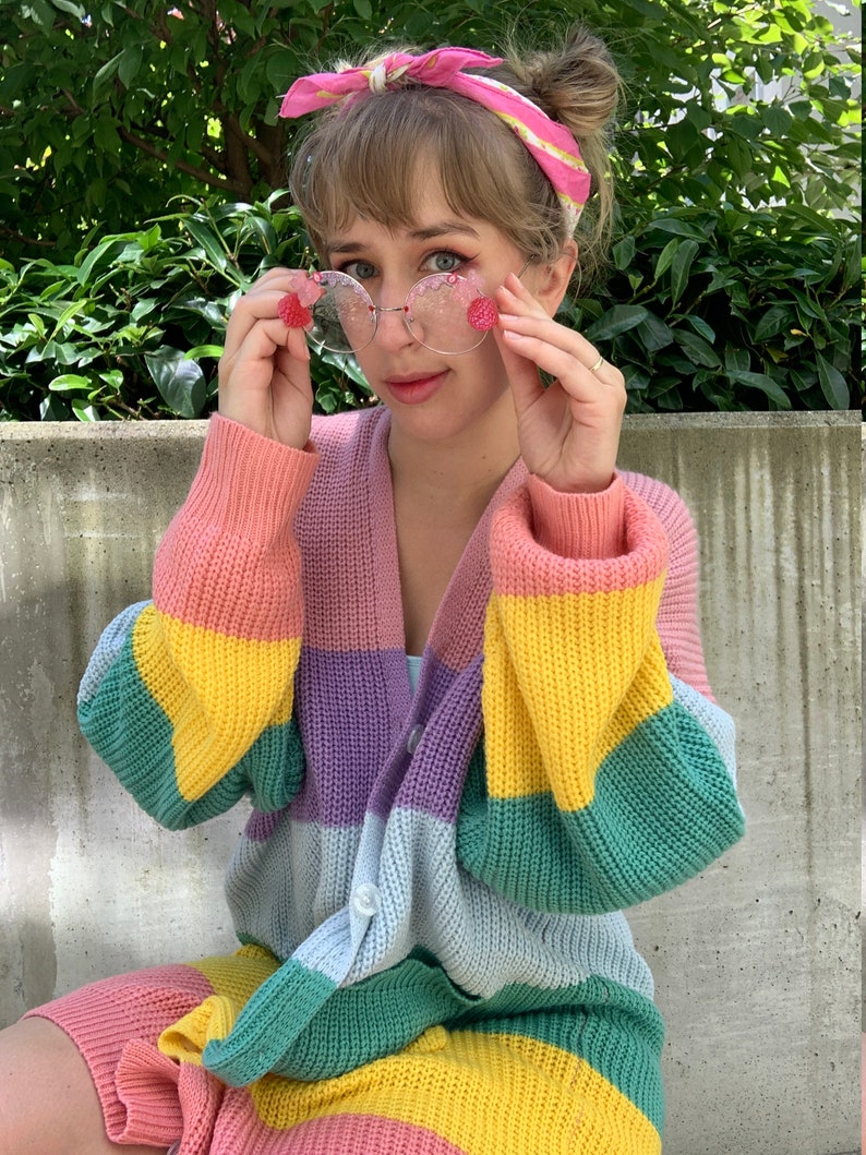 May include: A woman wearing a rainbow striped cardigan with a pink and yellow headband and round pink-tinted glasses.
