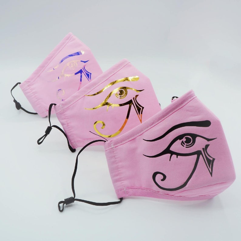 May include: Three pink face masks with an Egyptian eye design. The masks have black elastic ear loops. The eye designs are in gold, silver, and black.