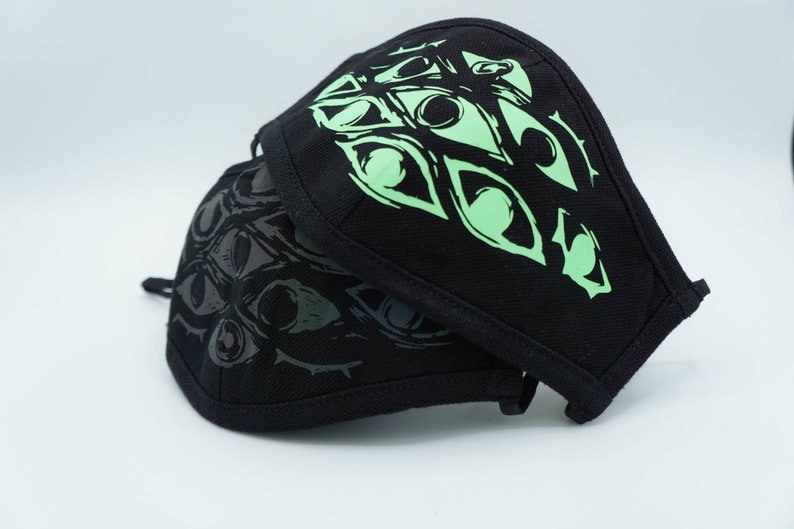 May include: Two black face masks with a repeating eye design. One mask has a light green eye design, and the other has a dark grey eye design. The masks are made of a fabric material and have a curved shape.