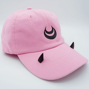 May include: Pink baseball cap with black crescent moon design and black horns on the brim.