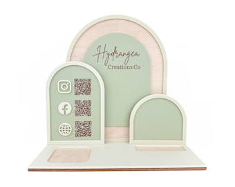 May include: A laser cut sign with a square reader slot, a business card holder, and a QR code for social media. The sign is made of wood and has a light green and brown color scheme. The text on the sign reads "Hydrangea Creations Co."