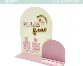 May include: A white and pink business card holder with a rounded arch top. The holder has a QR code, social media icons, and the text "Ellie Bean Embroidery".