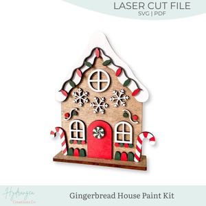 May include: A wooden gingerbread house paint kit featuring a laser cut design with a white roof, red door, two candy canes, snowflake cutouts, and windows. The text LASER CUT FILE SVG | PDF and Gingerbread House Paint Kit is visible.