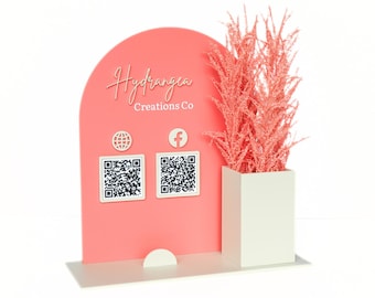 May include: A pink and white floral business card holder with a pink arch, the text "Hydrangea Creations Co", a globe icon, a Facebook icon, and two QR codes. The holder has a slot for business cards and a space for a potted plant.