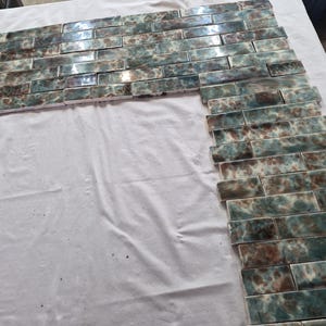 May include: Arrangement of rectangular ceramic tiles in shades of teal, brown, and white, resembling a brick pattern. The tiles are laid out on a white surface, creating a potential design for a wall or backsplash.