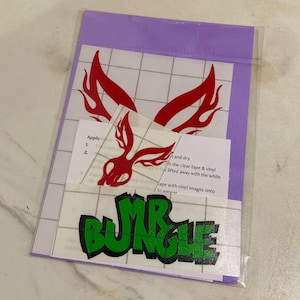 Puede incluir: Una calcomanía de vinilo roja y negra con llamas y el texto "MR BUNGLE" en letras verdes. La calcomanía está en una hoja de papel blanca con un patrón de cuadrícula.