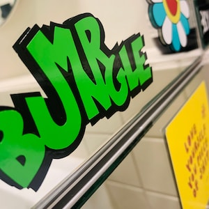 Puede incluir: Una pegatina verde y negra con la palabra "BUNGLE" escrita en una fuente de estilo graffiti.