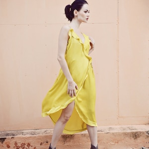 May include: A flowing, yellow wrap dress with ruffled trim and a V-neck. The dress is paired with brown ankle boots. The model is standing on a bed of small rocks in front of a peach-colored wall.