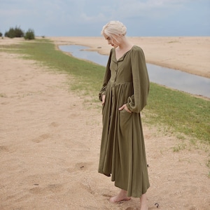 May include: A woman wearing a long, olive green, button-down dress with long sleeves and pockets. She is standing on a sandy beach with a small stream of water in the background.