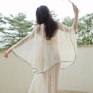 May include: A woman wearing a sheer white gown with a delicate gray trim. The gown is flowing and billowing in the breeze. She is holding a single pink rose in her hand.