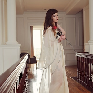 May include: A woman in a white sheer robe with a single red rose in her hand stands on a wooden floor in a hallway with a wooden railing.