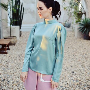 May include: A woman wearing a light blue, long-sleeved blouse with a high neckline and a pink skirt. The blouse has a slightly puffed sleeve and the skirt has a white pocket detail.