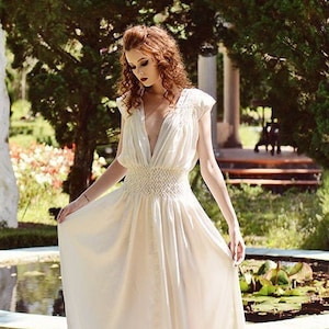 May include: A woman in a cream-colored, flowing gown with a deep V-neck stands in a garden. The dress has delicate lace detailing on the shoulders and a beaded waist. The garden features a pond, statues, and lush greenery.