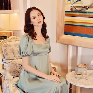 May include: A woman wearing a light green satin dress with short, puffed sleeves and a square neckline. She is seated on a vintage chair with a floral pattern. A small marble-topped table and artwork are in the background.