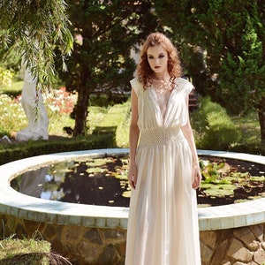 May include: A cream-colored, floor-length dress with a deep V-neck and a fitted waist. The dress features a flowing skirt and cap sleeves. The woman is standing in a garden with a pond in the background.