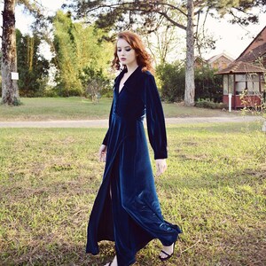 May include: A long, dark teal velvet dress with long sleeves and a high slit. The dress is buttoned at the waist and falls to the ankles. The person is wearing black sandals and walking on grass.
