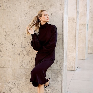 May include: A woman wearing a black turtleneck dress with a long sleeve and a fitted silhouette. She is standing next to a concrete wall and wearing black heels with a jeweled buckle.