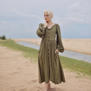 May include: A woman wearing a long-sleeved, olive green button-down dress with a gathered skirt and puffed sleeves. She is standing on a sandy beach with a small stream of water in the background.