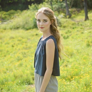 May include: A woman with long blonde hair wearing a dark blue sleeveless top and light gray pants stands in a field of yellow wildflowers.