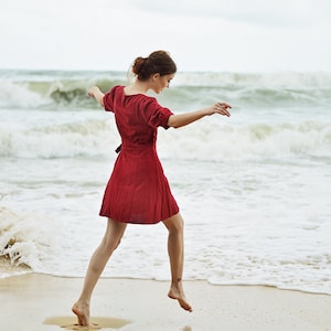 May include: A woman in a red dress jumps in the air on a sandy beach with waves crashing in the background.