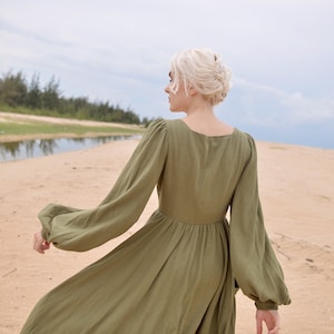 May include: A woman in a long, olive green dress with puffed sleeves walks on a sandy beach. The dress has a square neckline and a flowing skirt.