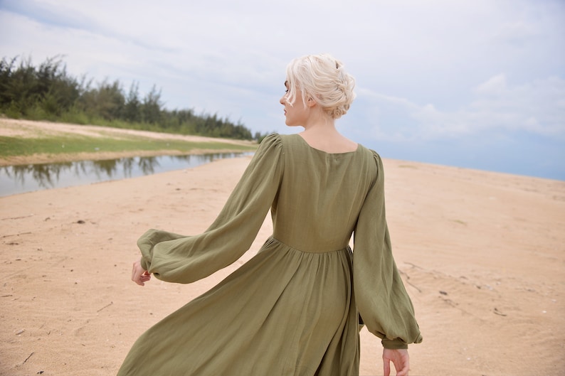 May include: A woman in a long, olive green dress with puffed sleeves walks on a sandy beach. The dress has a square neckline and a flowing skirt.