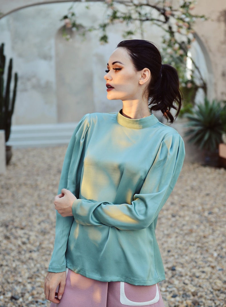 May include: A woman wearing a teal satin blouse with long sleeves and a high neckline. She is wearing a pink skirt with a white trim.
