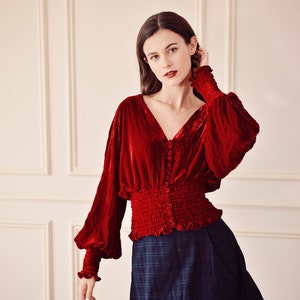 May include: A red velvet blouse with long sleeves and a button-up front. The blouse has a gathered waist and a ruffled hem. The model is wearing a dark blue plaid skirt.