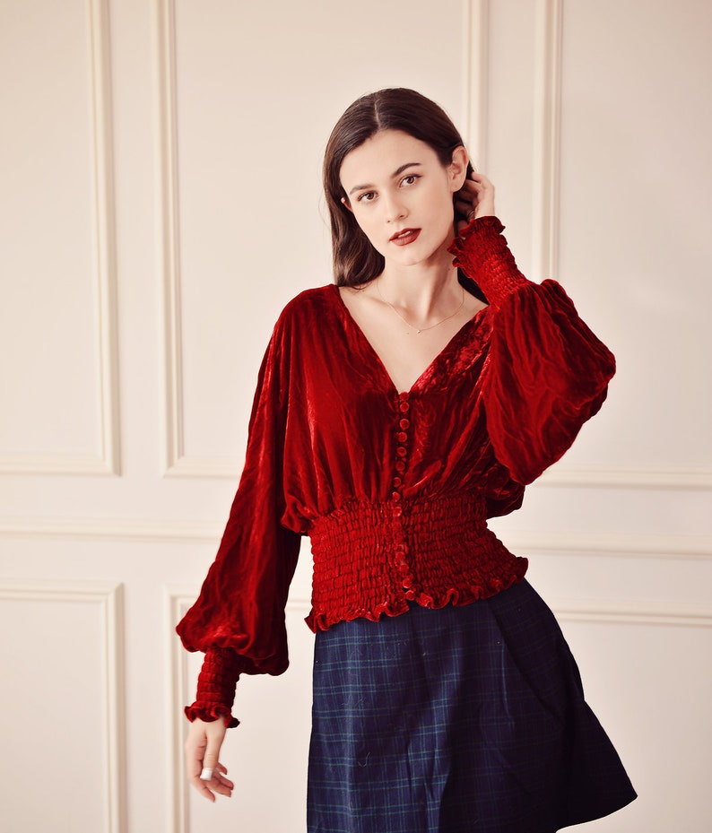 May include: A red velvet blouse with long sleeves and a button-up front. The blouse has a gathered waist and a ruffled hem. The model is wearing a dark blue plaid skirt.