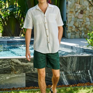 May include: A man wearing a white linen shirt with a button-down collar and a dark green pair of shorts. He is standing in front of a swimming pool and a stone wall.