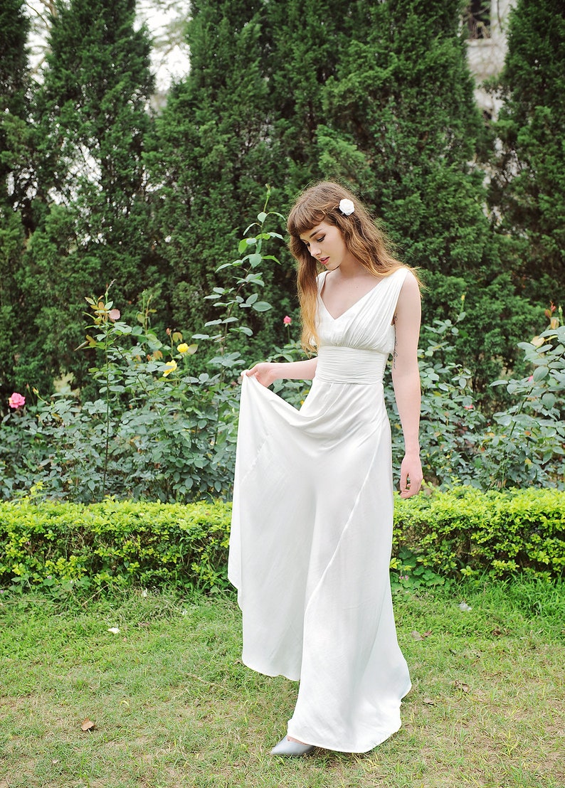 May include: A woman in a white sleeveless dress with a V-neckline and a sash at the waist stands in a garden setting. She is holding the skirt of the dress in her left hand and looking down.
