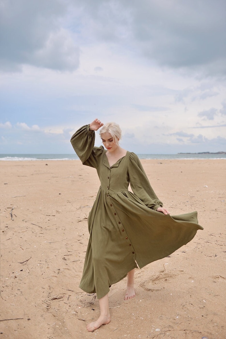 May include: A woman in a long, olive green, button-down dress with puffed sleeves stands on a sandy beach. The dress has a flowing skirt and the woman is barefoot.
