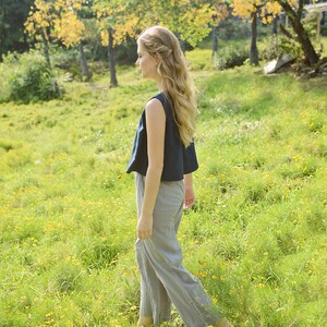 May include: A woman with long, wavy blonde hair walks through a field of green grass and yellow wildflowers. She is wearing a dark blue sleeveless top and gray pants.